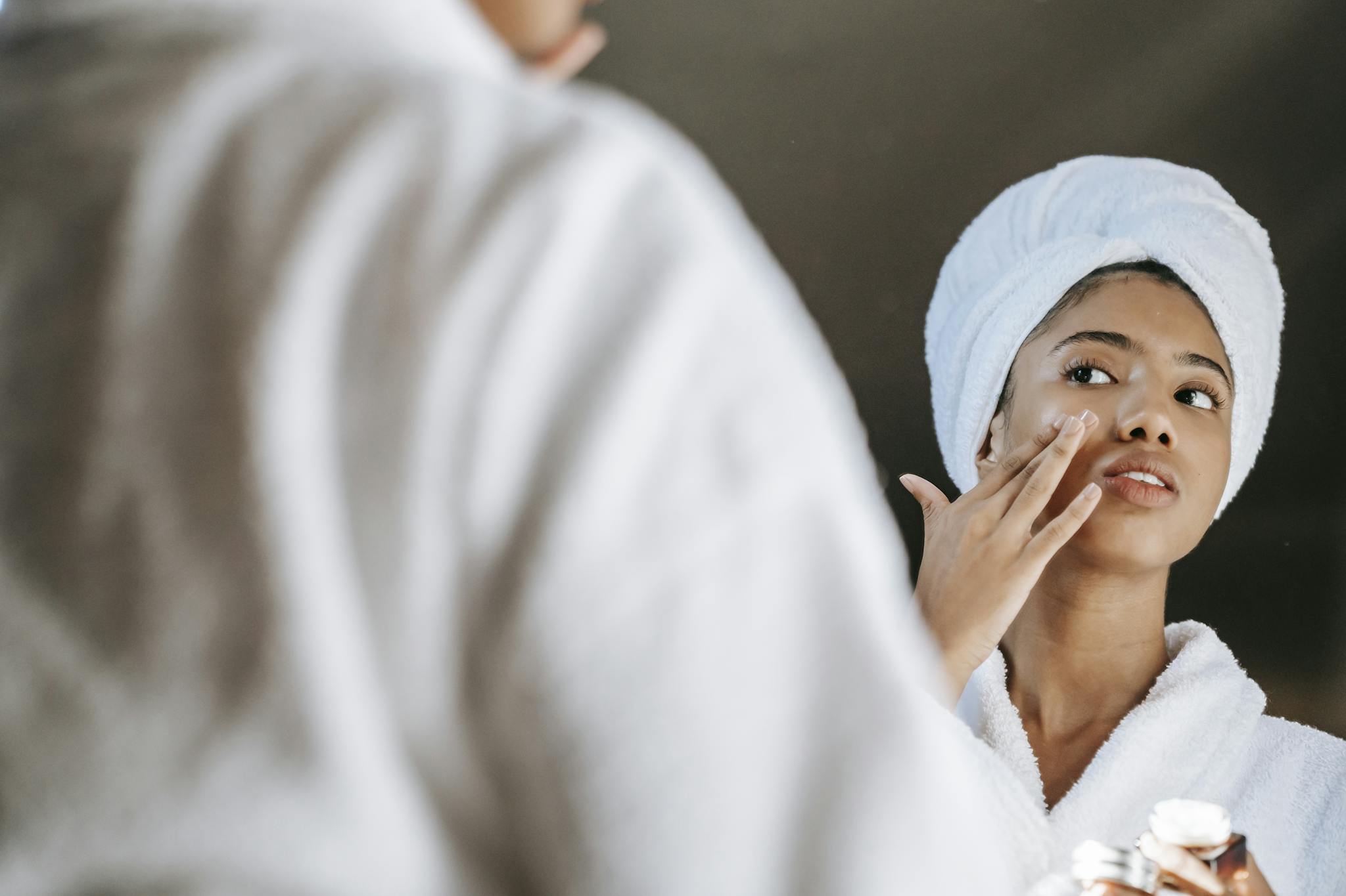 A young woman applies skincare cream while wearing a bathrobe in front of a mirror.