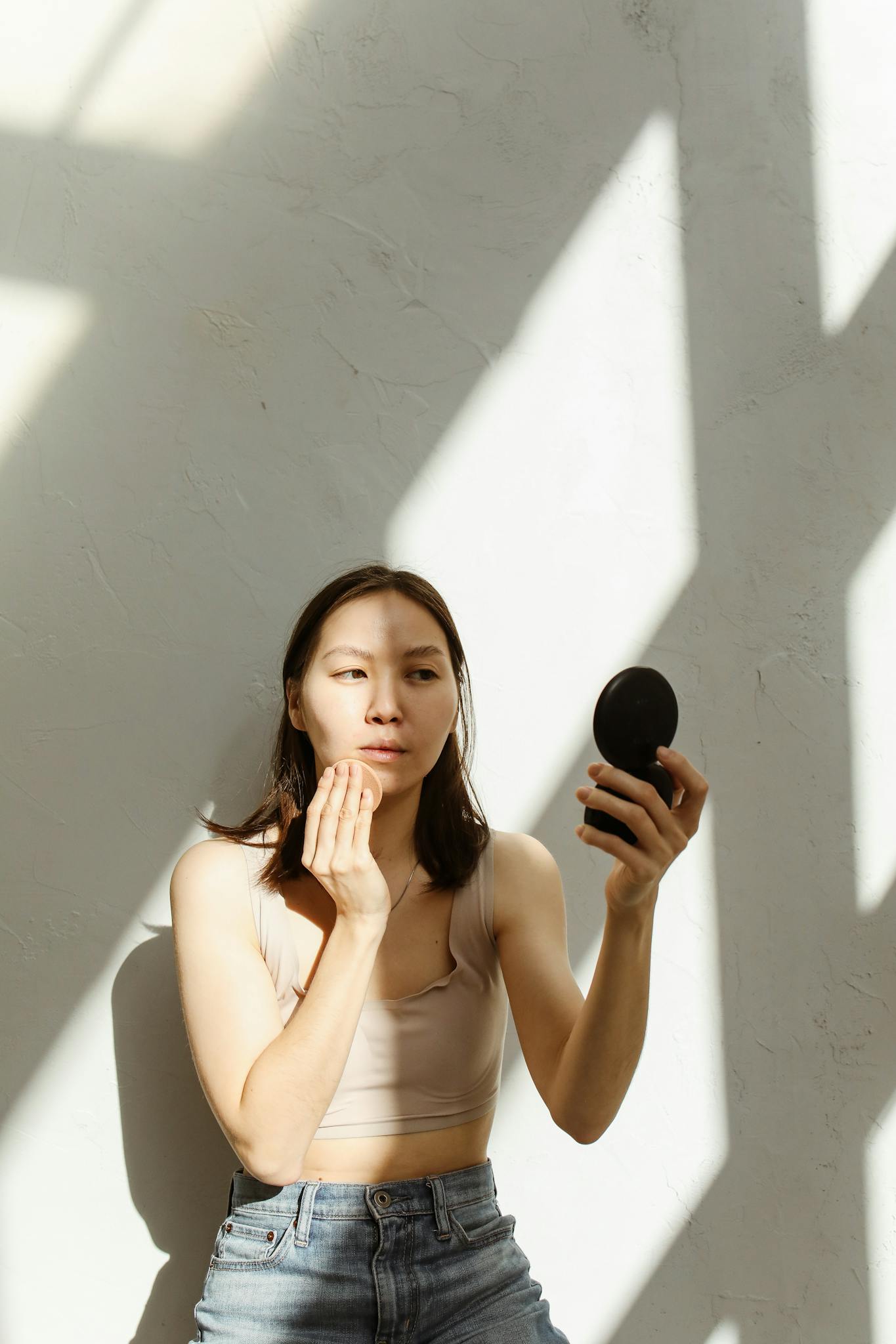 Asian woman applying makeup while sitting indoors, illuminated by natural light and shadows.